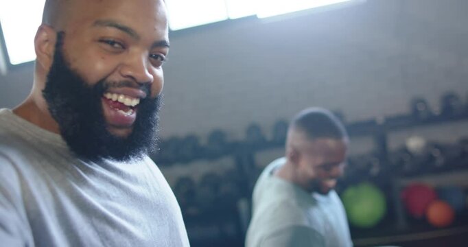 African American men laughing, encouraging partner curling hex dumbbell in gym in workout shirts