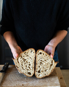 Close-up of hands holding a sourdough bread loaf cut in half on a wooden cutting board, showing airy open crumb and crust.