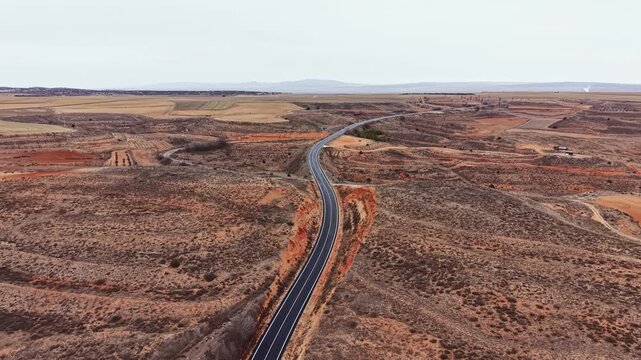 A long road curves through open countryside with fields and flat terrain. The sky is gray and the landscape appears dry with patches of land visible.