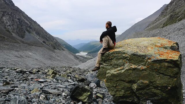 A guy sits on a huge rock in the mountains and admires the river valley