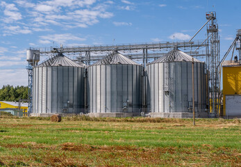 Obraz premium Agro-processing and manufacturing plant with silver silos for drying, cleaning, and storage of agricultural products. Grain elevator complex in a rural field with hay bales in the foreground.