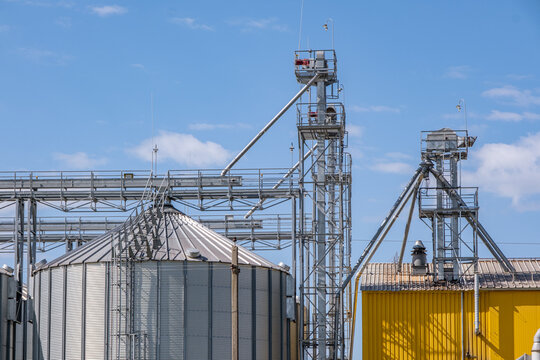 Modern agro-processing plant with silver metal silos for grain storage. Industrial granary elevator complex for drying and cleaning agricultural products against a blue cloudy sky.
