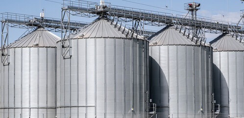 Industrial grain elevator and agro-processing plant with metal silos and conveyor system. Agricultural facility for drying, cleaning, and storage of crops against a blue cloudy sky. © NastyaPhoto