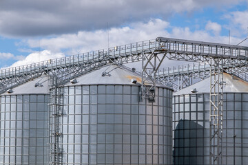 Fototapeta premium Industrial grain elevator and agro-processing plant with metal silos and conveyor system. Agricultural facility for drying, cleaning, and storage of crops against a blue cloudy sky.
