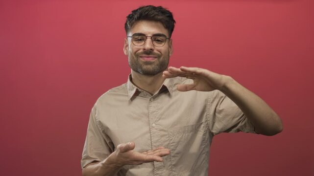 Man with hands framing imaginary object in a red studio wearing round glasses and beige shirt; confidence.