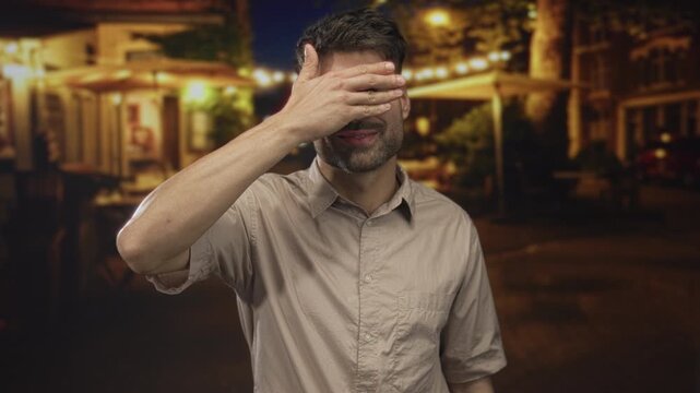 Man covers eyes with hand on deserted city street at night under glowing streetlights by cafe facades; shame.