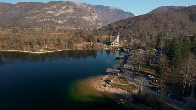 Lake Bohinj Slovenia St John the Baptist church drone reveal. Dark water pine reflections, Julian Alps mountains landscape. Moody autumn travel 4K video.