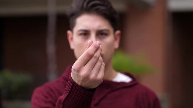 Young man wearing a burgundy sweater making a money gesture with his hand, expressing the need for cash, financial pressure, and economic concern.