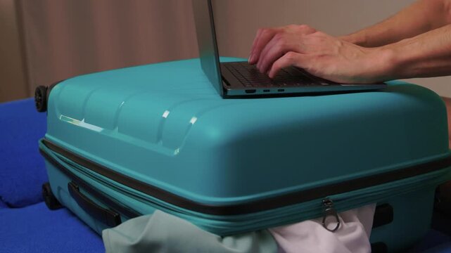 Laptop on turquoise suitcase, hands typing on keyboard on closed case improvised workspace in hotel room with clothes peeking from zip, soft evening light, focused productive mood
