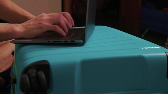 Laptop on turquoise suitcase, hands typing on keyboard on closed case improvised workspace in hotel room with clothes peeking from zip, soft evening light, focused productive mood