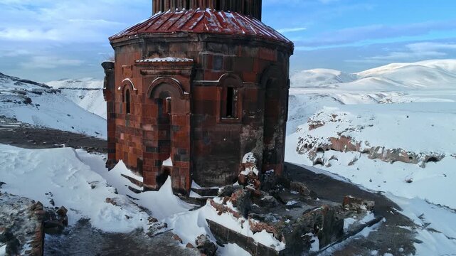 Close aerial view of an ancient Armenian church at Ani Ruins in Kars, Turkey. Snow covered red stone medieval architecture stands against a dramatic winter landscape.