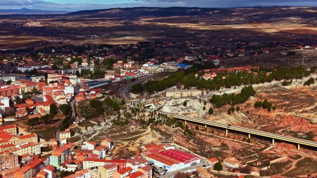 The town shows houses and buildings scattered along the valley. Roads connect different areas. Hills and open land are visible in the background under a clear sky.
