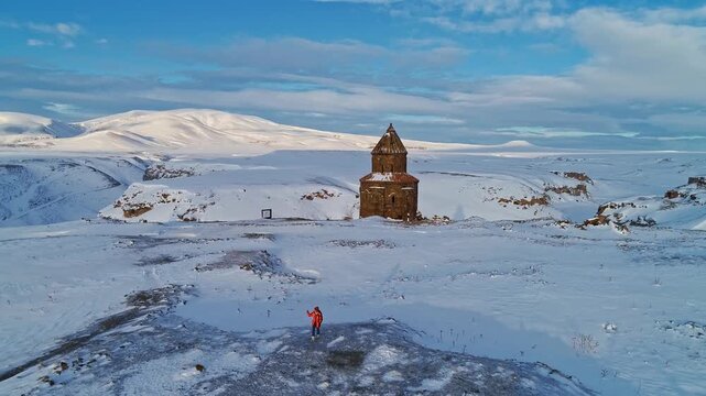 Aerial view of a tourist photographing the Abughamrents Church at Ani Ruins in Kars, Turkey. Snow covered medieval Armenian chapel stands in a vast winter landscape.