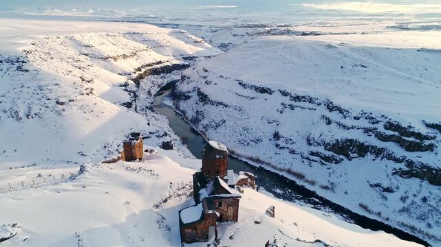 Aerial view of the Menucehr Mosque minaret at Ani Ruins overlooking a snowy canyon in Kars, Turkey. Medieval stone architecture rises above a dramatic winter landscape.