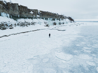 Aerial drone view of a lonely man walking on frozen sea ice near high limestone cliffs with icicles in Paldiski.