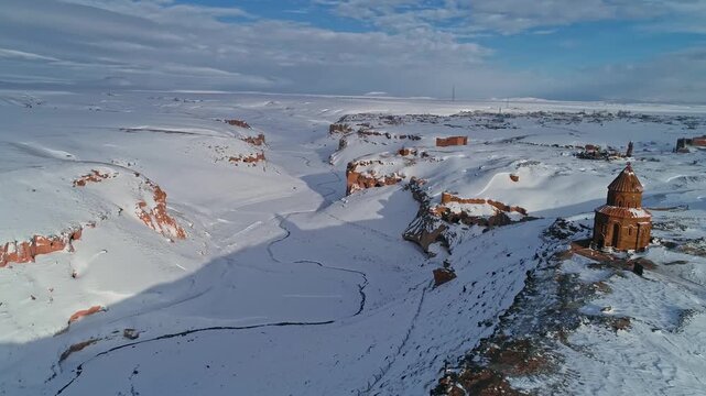 Wide aerial view of Ani Ruins in Kars, Turkey. Snow covered medieval structures stand within a vast Anatolian winter landscape under a dramatic cloudy sky.