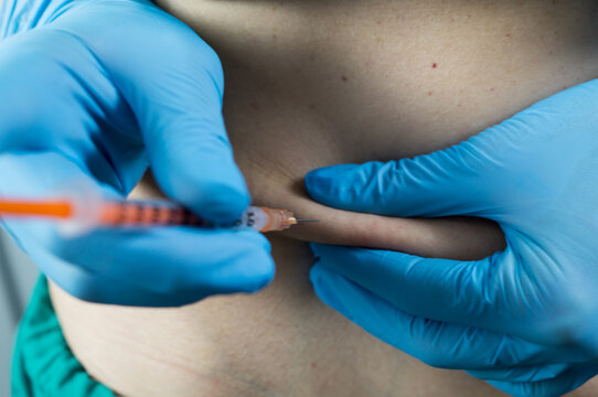 Close-up of a medical professional's hands in blue gloves pinching skin on the belly to administer a subcutaneous injection, such as insulin or heparin.