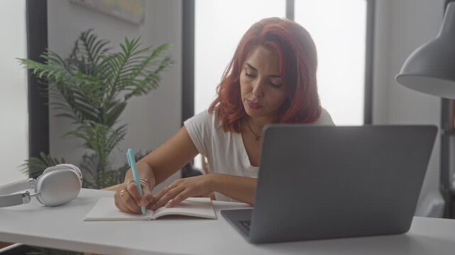 Woman writing in notebook with turquoise pen and laptop at desk in building; concentration productivity.