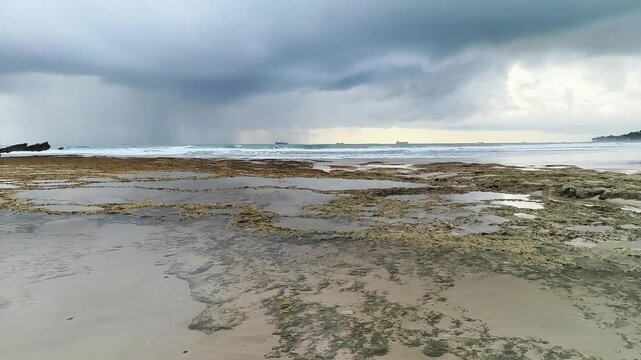 Dramatic storm clouds over Bayah Beach near Sawarna, Indonesia. Rocky coastline at low tide with crashing waves, dark rainy sky, and ships on the horizon. Moody tropical seascape in Banten.