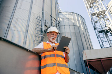 Senior Male Industry Worker Using Tablet Outdoors At Industrial Facility With Silos