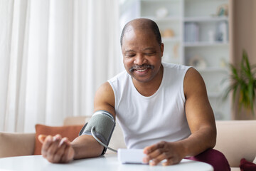 Mature black man checking blood pressure at home with digital monitor, smiling calmly. Domestic lifestyle scene showing health control and preventive care concept