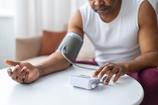 Mature black man measuring blood pressure at home using digital monitor while sitting at table. Domestic lifestyle scene highlighting health control and hypertension awareness
