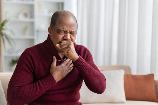 Mature black man coughing with hand on chest while sitting on sofa at home. Domestic lifestyle scene showing respiratory illness, cold symptoms and healthcare concept
