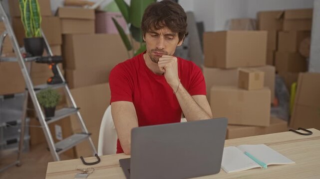 Man resting chin on fist while seated at laptop surrounded by cardboard boxes in building; quiet contemplation.