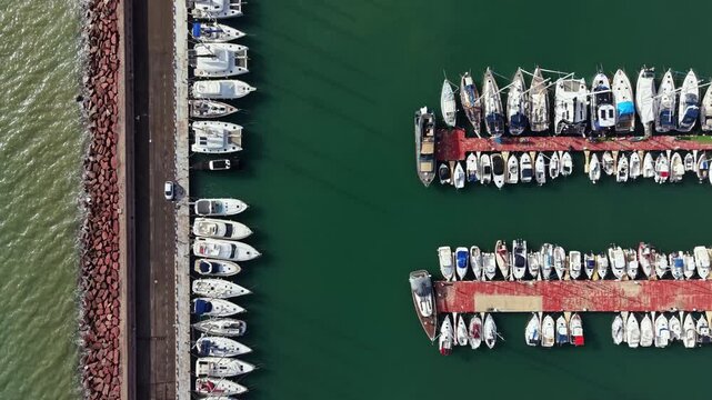 Boats are lined up in neat rows at a marina. Docks are visible with boats tied to them. The water reflects the colors of the boats. A road runs parallel to the marina.