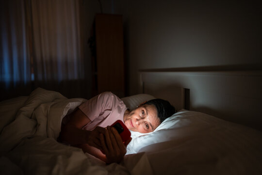 Sleepy senior woman phone addict scrolling social media online on smartphone, lady lying in bed in darkness looking at bright phone screen, unhealthy habit leading to eyesight problems and insomnia.