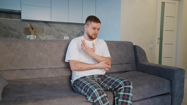 Young man in pajamas scratching an itchy arm at home