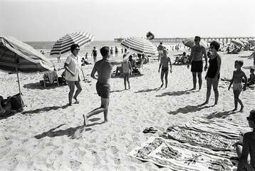 A black and white vintage photograph of a group of people playing volleyball on a sunny, crowded beach in the 1960s. © Stitch