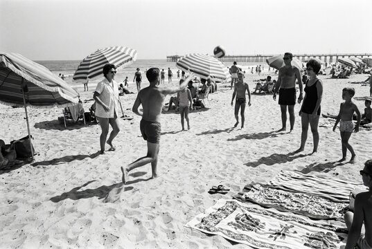 A black and white vintage photograph of a group of people playing volleyball on a sunny, crowded beach in the 1960s.