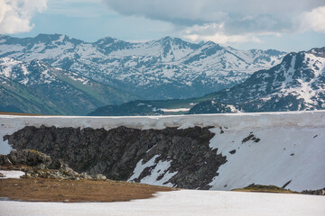 Scenic view from sunlit snow field to gold stone outcrops and sharp golden rocks against pointy peaks and peaked tops under cloudy sky. Wild rugged alpine landscape with big rocky snowy mountain range © Daniil