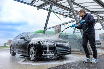 Man washing black car with high pressure washer at self service car wash station outdoors. A man cleans a black car using a high pressure washer at a self service car wash station.  © Diatlova Tetiana