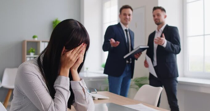 Woman stressed by workplace bullying and gossip. A distressed employee hides her face at the desk as coworkers chat, depicting workplace harassment, stress, anxiety, sadness and toxic culture.