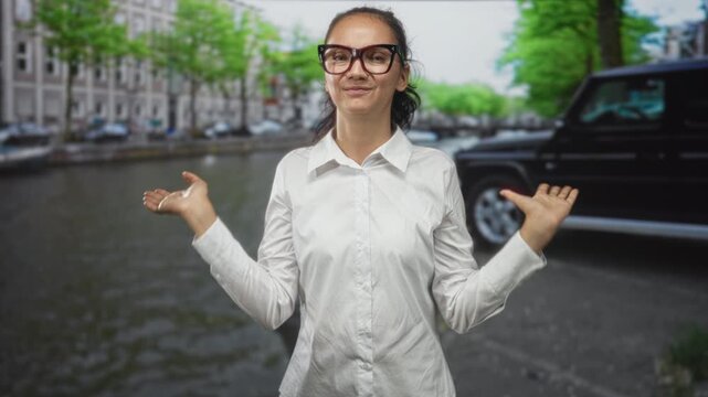 Woman wearing glasses shrugs with palms up beside canal on a city street near parked suv and row of buildings; casual confidence.