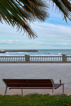 Wooden bench and palm leaves framing Mediterranean seafront promenade, tranquil coastal scene with ocean waves and balustrade