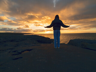 Silhouette of a woman standing on stone coast and looking at amazing sunset. Burren, Ireland. Travel and tourism. Hand up pose. Embrace nature beauty concept.