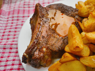 A plate of food with a fork and knife on a checkered cloth. The plate has meat and potatoes and cabbage salad. Dinner with excellent beef on the bone and wedges. Premium product.