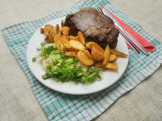 A plate of food with a fork and knife on a checkered cloth. The plate has meat and potatoes and cabbage salad. Dinner with excellent beef on the bone and wedges. Premium product.