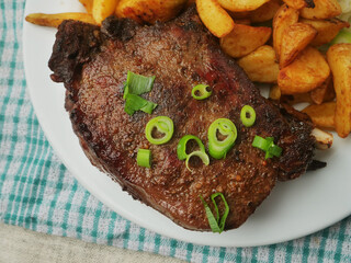A plate of food with a fork and knife on a checkered cloth. The plate has meat and potatoes and cabbage salad. Dinner with excellent beef on the bone and wedges. Premium product.