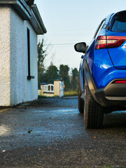 A blue car is parked in front of a white house. The car is parked on a wet road, and the reflection of the car can be seen in the water. The scene is calm and peaceful. Rural country side area.