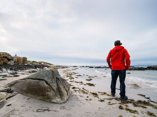A man in a red jacket is standing by a rock on a sandy beach. The sky is cloudy and the beach is empty. Cloudy sky. West coast of Ireland. Travel and tourism. Outdoor activity
