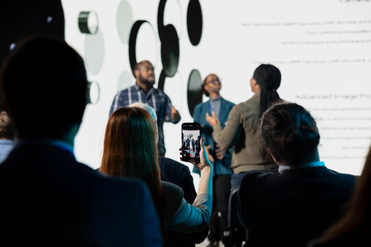 Selective focus of young woman streaming live from product launch at business convention, creating content while being in attendance for career growth. Attendee takes photos of new phone.
