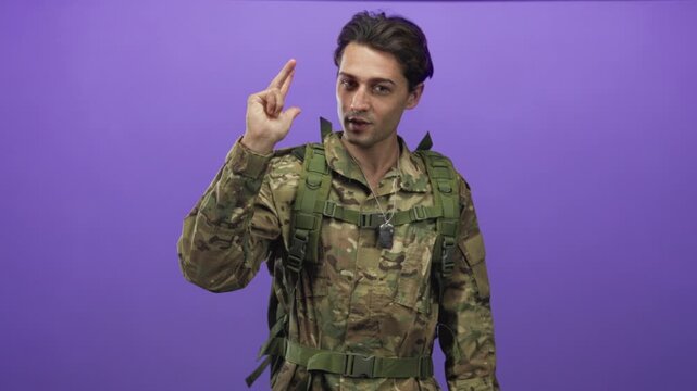Man in camouflage military uniform with dogtag necklace and tactical backpack raises two fingers in a finger gun gesture near face in studio purple backdrop; duty readiness.