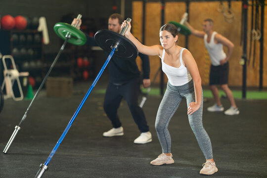 Determined sporty young girl doing one arm landmine press with barbell in high-intensity group training session at modern gym, focusing on shoulder and core muscles....