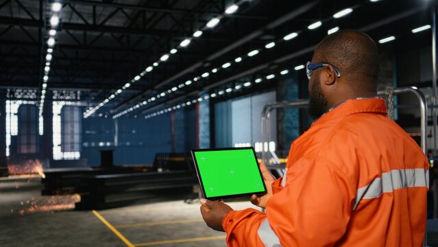 Green screen tablet with worker managing operation tasks across the factory floor, surrounded by metal machinery and fabrication tools that highlight professional engineering.