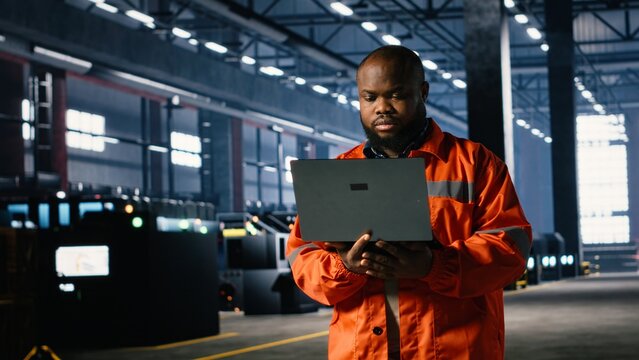 Industrial staff member oversees assembly work in a warehouse while using laptop to support professional supervision and ensure safe and efficient steel manufacture and production flow.