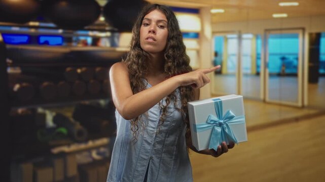 Teenage woman holding gift box with blue ribbon points finger to the side in studio; confidence offering.